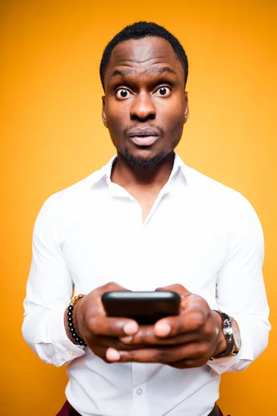 Όμορφος Αφροαμερικάνος που κρατάει ένα τηλέφωνο στο χέρι του και εκπλήσσεται από τη δύναμή του ενώ στέκεται σε κίτρινο φόντο — Φωτογραφία Αρχείου