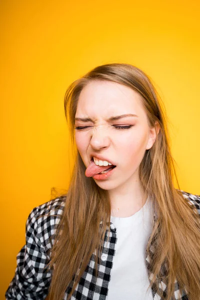 Uma menina bonita fica em um fundo amarelo e se destaca sua língua para o lado, fechando os olhos — Fotografia de Stock