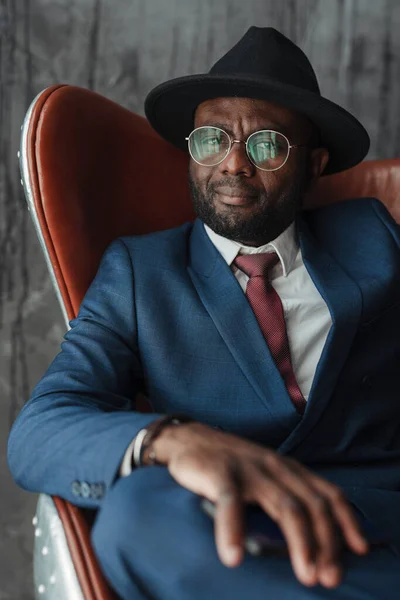 Serious stylish african american businessman in a hat with glasses and in a suit sits on a chair and looks to the side — Φωτογραφία Αρχείου