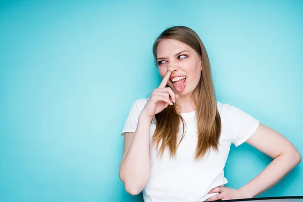 Roztomilá veselá dívka v bílém tričku se prstem dotkne nosu a vystrčí jazyk — Stock fotografie