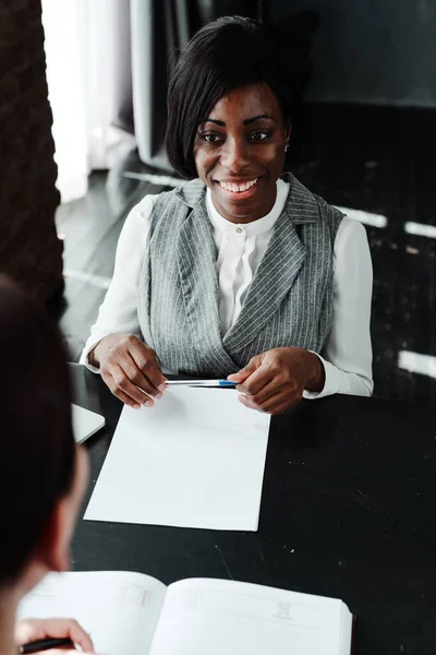 Χαρούμενη Αφροαμερικανή που κάθεται στο τραπέζι και παίρνει συνέντευξη κρατώντας στυλό στο χέρι. — Φωτογραφία Αρχείου