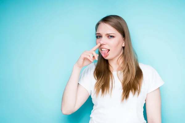 Une belle fille mignonne dans un T-shirt blanc touche son nez avec son doigt et sort sa langue grimaçante d'une mauvaise odeur — Photo