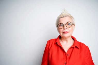 Gözlüklü, kırmızı tişörtlü, gri arka planda kameraya bakan güzel, yaşlı bir kadın.