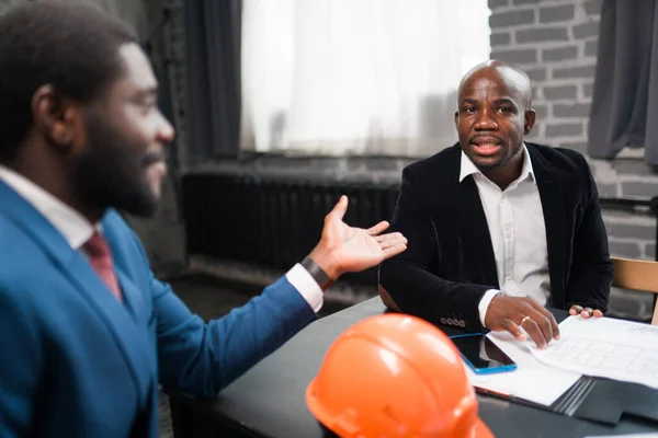 Два афроамериканских бизнесмена сидят за столом в строительном шлеме и ведут переговоры — стоковое фото