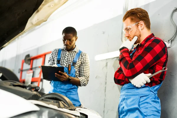 Afroameričtí a rusovlasí řemeslníci stojí v montérkách nad otevřenou kapotou auta, definují problém a přemýšlejí — Stock fotografie