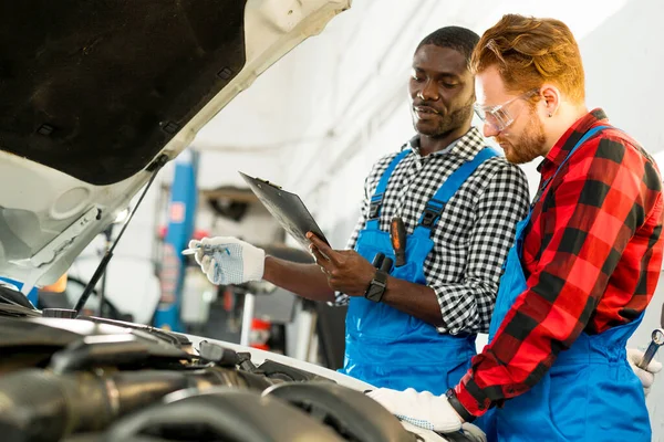 Africký americký mechanik učí mladého chlapa o automobilových součástech v autoservisu, zatímco stojí nad otevřenou kapotou — Stock fotografie