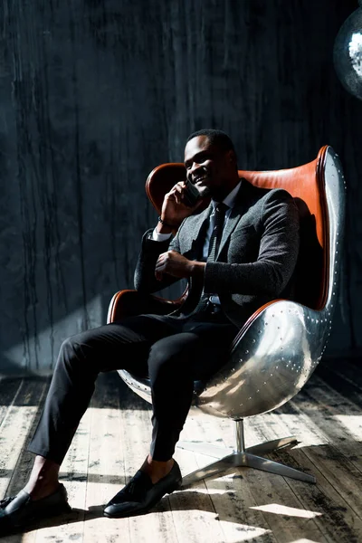 Bonito empresário afro-americano com uma jaqueta e calças senta-se em uma cadeira e olha para o lado sorrindo — Fotografia de Stock