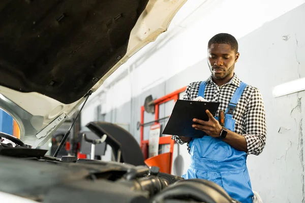 Africano americano mecânico de automóveis homem em macacão e segurando um caderno em suas mãos, enquanto está perto do capô aberto do carro — Fotografia de Stock