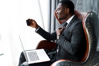 Yakışıklı Afro-Amerikan iş adamı bir sandalyede oturuyor ve dizüstü bilgisayarda çalışıyor ve elinde bir telefon tutuyor.