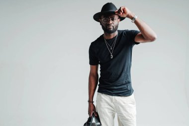 Şapkalı yakışıklı Afro-Amerikan adam kameraya bakıp elinde bir çanta tutarken elini tutuyor.