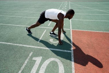 Yandan bir fotoğraf. Tişörtlü ve şortlu Afro-Amerikalı bir sporcu yarışa hazırlanırken...