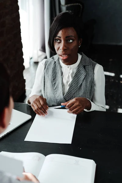Переживающая африканская американка, сидящая за столом и проводящая собеседование, держащая ручку — стоковое фото