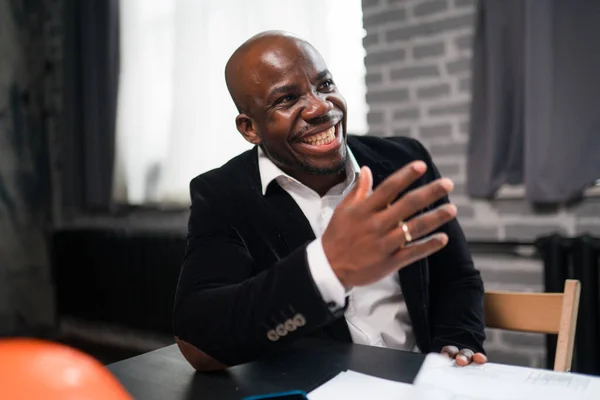 Feliz empresário afro-americano rindo e gesticulando enquanto se senta à mesa e conversa com colegas — Fotografia de Stock