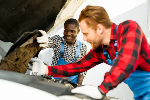 African American and red-haired auto mechanics in overalls check the engine oil level in the open hood of the car — Stock Photo, Image