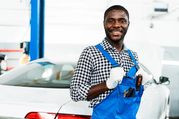 Šťastný africký americký automechanik muž v montérkách stojí na pozadí auta v prodejně aut — Stock fotografie