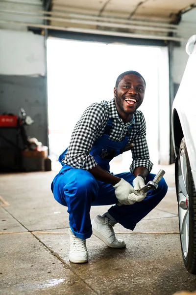 Happy african american mechanic man sitting next to a wheel of a car and repairing it