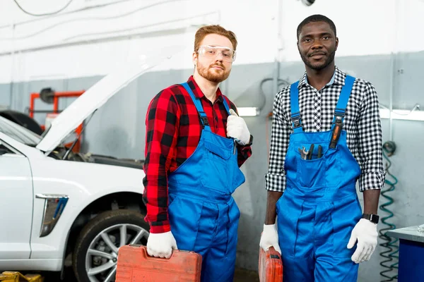 Afroameričtí a rusovlasí mechanici drží kufr s nářadím v rukou a stojí na pozadí auta — Stock fotografie