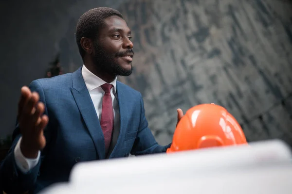 Όμορφος αφροαμερικάνος επιχειρηματίας μηχανικός που μιλάει ενώ κάθεται στο τραπέζι και με ένα κράνος που βρίσκεται πάνω του — Φωτογραφία Αρχείου