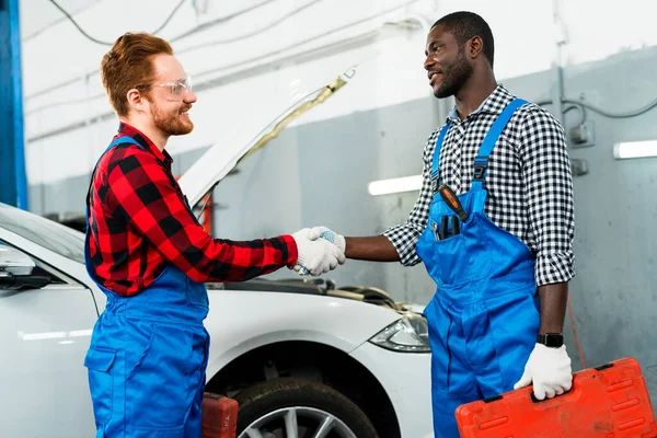 Africký Američan drží nářadí a červenovlasý mechanik potřásá rukou, zatímco stojí na pozadí auta v autoservisu — Stock fotografie