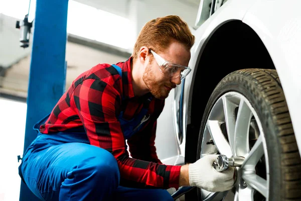 Muž mechanik v brýlích a v montérkách mění pneumatiku na automobilovém kole v autoservisu pomocí klíče — Stock fotografie