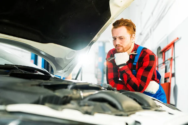 Seriózní automechanik v kostkované košili a montérkách přemýšlí o problému s autem, zatímco drží klíč v rukou a stojí u otevřené kapoty — Stock fotografie