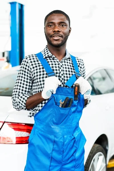 Hezký africký americký samec automechanik v montérkách a rukavicích stojí v autosalonu na pozadí auta — Stock fotografie