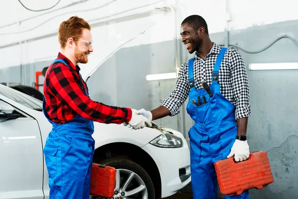 Afroameričtí a rusovlasí mladí mechanici v montérkách si potřásají rukou, když stojí v prodejně aut — Stock fotografie