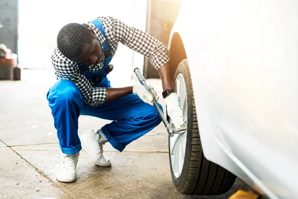 Afroameričan auto mechanik sedí v blízkosti kola auta a nahrazuje pneumatiku, zatímco drží klíč v ruce — Stock fotografie