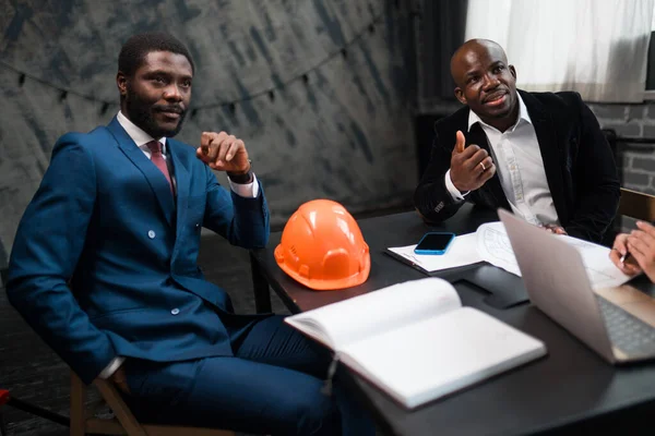 Dois empresários afro-americanos estão sentados em uma mesa de negociação com um capacete de construção deitado sobre a mesa — Fotografia de Stock