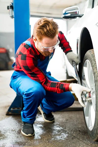 Muž automechanik v brýlích a montérkách zkontroluje kolo auta a nahradí pneumatiku klíčem — Stock fotografie