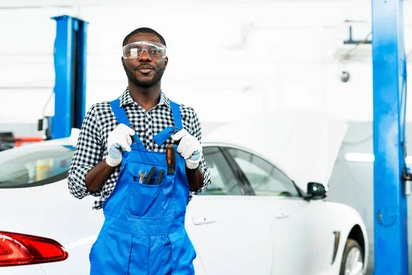 Sebevědomý afro americký mechanik v ochraně brýlí a modrého ptáka s bílým autem vzadu připraven k práci. — Stock fotografie