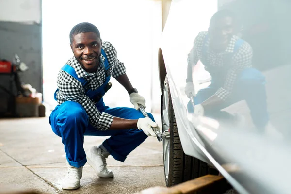 Afro americký mechanik drží klíč v rukou a odšroubovat pneumatiku auta v modrém dickey. — Stock fotografie