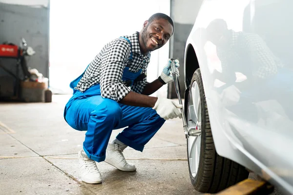 Šťastný africký mechanik s ráčkovým klíčem opravující pneumatiku v bílých rukavicích a modrém péru. Výměna pneumatiky — Stock fotografie