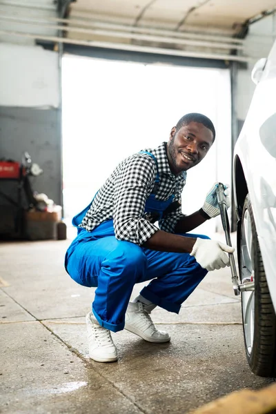 Šťastný africký mechanik s ráčkovým klíčem opravující pneumatiku v bílých rukavicích a modrých gumových botách. Výměna pneumatiky — Stock fotografie
