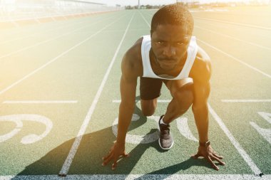 Koyu tenli erkek bir atlet ciddi bir yüzle başlangıç çizgisinde duruyor ve ellerine yaslanıyor.