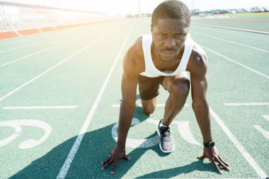 Koyu tenli erkek bir atlet ciddi bir yüzle başlangıç çizgisinde duruyor ve ellerine yaslanıyor.
