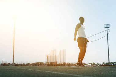 Beyaz tişörtlü siyah bir erkek atlet kavurucu güneşin arka planına karşı bir iple idman yapıyor.