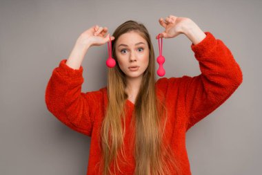 Kırmızı kazaklı, beyaz tenli çekici bir kız şaşkınlığın heyecanını anlatıyor. Ağzını açıyor ve elinde kırmızı vajinal toplar tutuyor. Seks için masaj aleti.