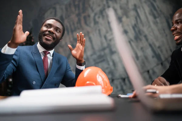 Ingeniero empresario afroamericano hablando mientras está sentado en la mesa y con un casco de construcción acostado en él — Foto de Stock