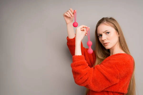 Una mujer con bolas de Kegel vaginal de color rojo brillante en sus manos se ve con una cara reflexiva sobre un fondo gris —  Fotos de Stock