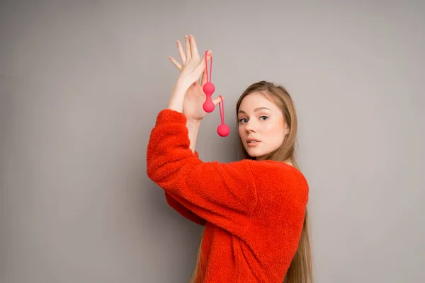 Een vrouw met felrode vaginale Kegel ballen in haar handen maakt een verrassende blik, geïsoleerd op een grijze achtergrond Rechtenvrije Stockfoto's