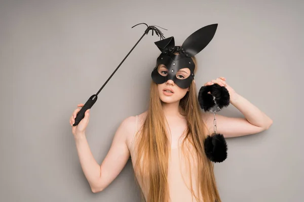 Een meisje in een zwart konijn leer fetisj masker met handboeien in haar handen, een zweep voor seks op een grijze achtergrond Stockfoto