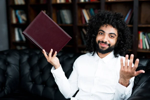 An exchange student spreads his hands to the side, holding a book in the library against the background of books