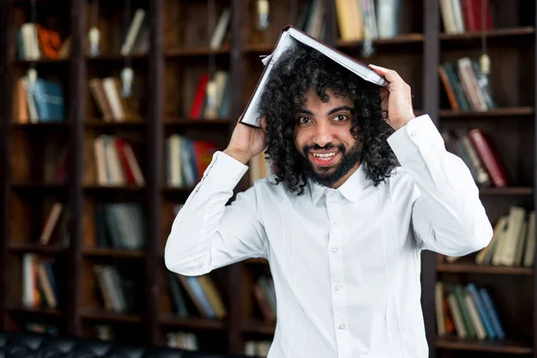 Ein breit lächelnder ägyptischer Student hält in der Bibliothek vor dem Hintergrund von Büchern ein Buch über dem Kopf lizenzfreie Stockbilder