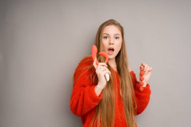 An attractive girl in a sweater is surprised, opening her mouth, holding a red dildo,vaginal balls in her hand