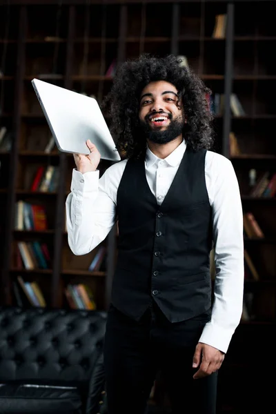 Een leuke student uit Turkije houdt een laptop in zijn handen en lacht breed tegen de achtergrond van boeken Rechtenvrije Stockfoto's