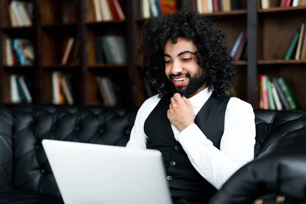 Un estudiante afroamericano en un traje se dedica en línea con un tutor en un ordenador portátil en el fondo de los libros Imagen De Stock