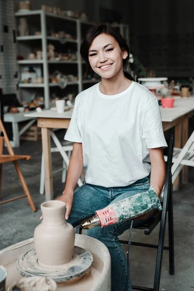 Schattig jong aziatisch meisje in een t-shirt en jeans zit op een stoel het maken van een klei vaas op een pottenbakkers whee — Stockfoto