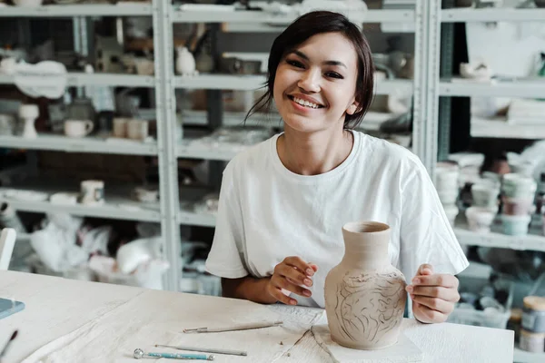 Een aantrekkelijk Aziatisch meisje in een wit T-shirt zit aan een tafel en glimlacht prachtig in een pottenbakkerij en houdt een zelfgemaakte kleivaas in haar hand. Hobby studio concept. — Stockfoto