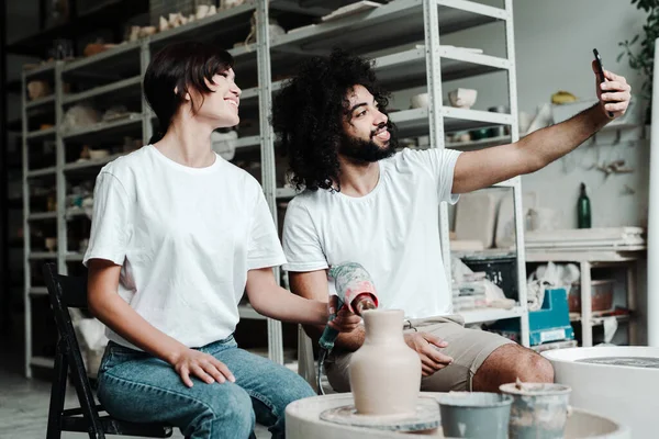 In einer Kreativwerkstatt machen ein verliebtes türkisches und ein asiatisches Paar ein Selfie. Herstellung einer Tonvase. Stockbild